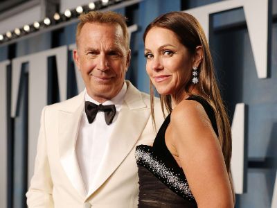 Kevin Costner is wearing a white suit with black bow tie and Christine Baumgartner is wearing a black dress.